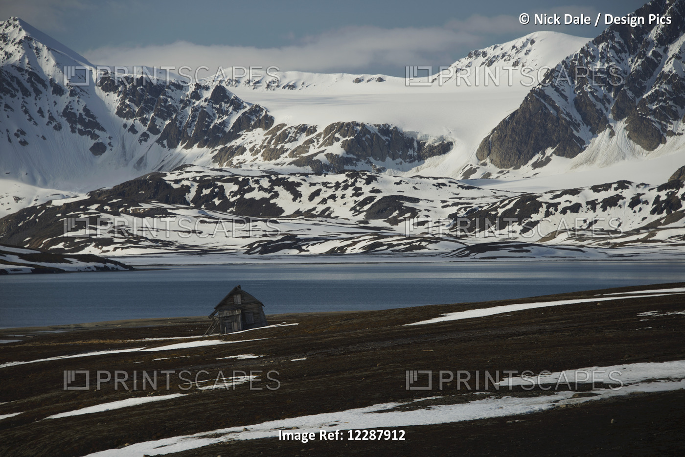 Small Wooden Cabin On The Shore Of The Arctic Ocean And Snow Covered Mountains; ...