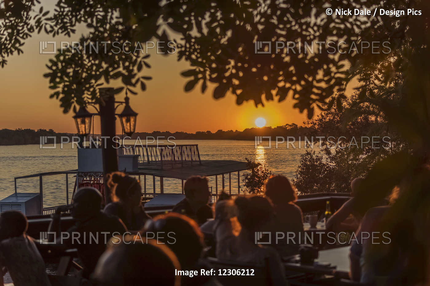 People Sitting On A Patio, Backlit By The Setting Sun And Framed By Tree ...