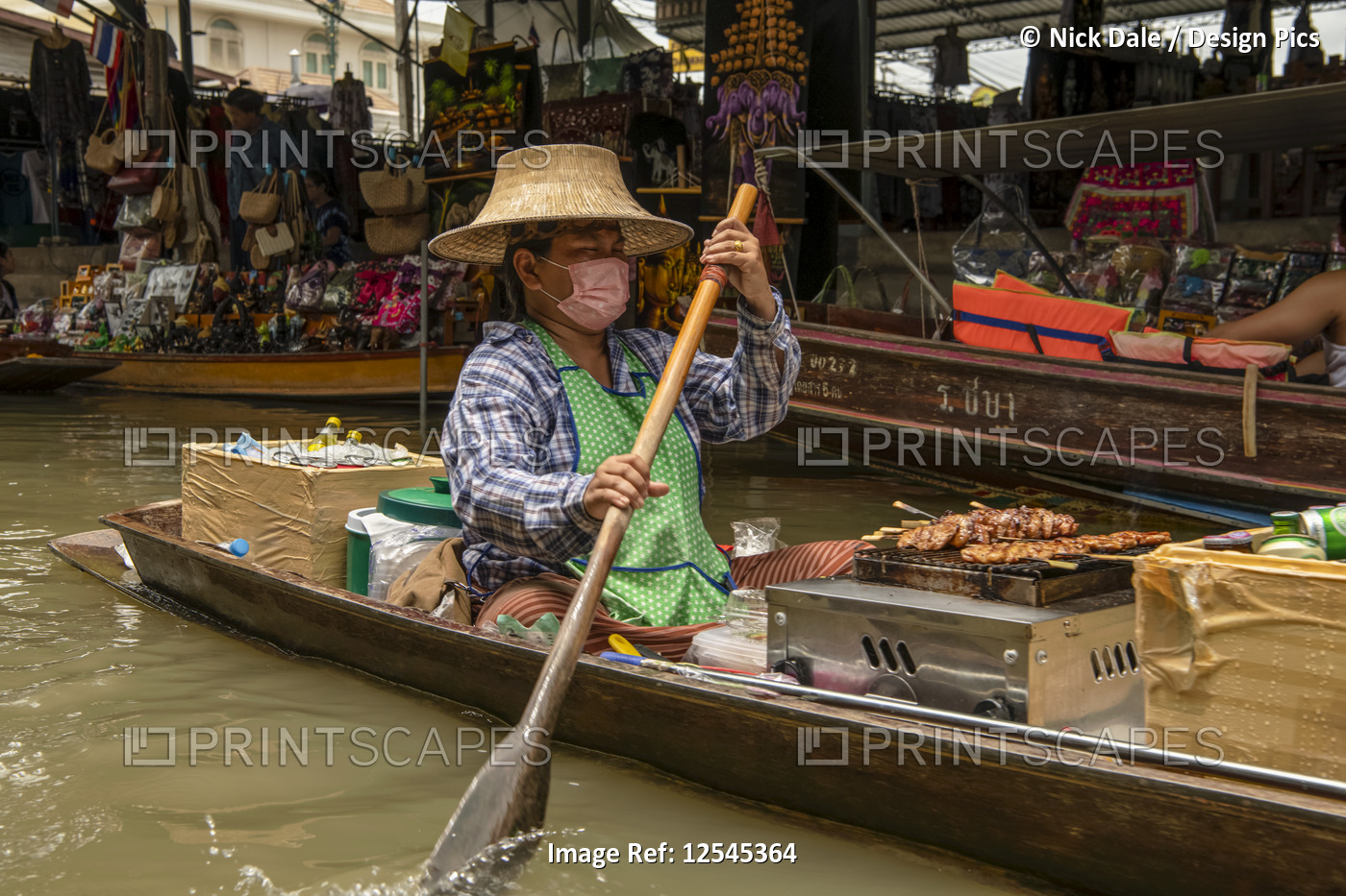 Man with mask paddles through floating market with skewers on a grill on his ...